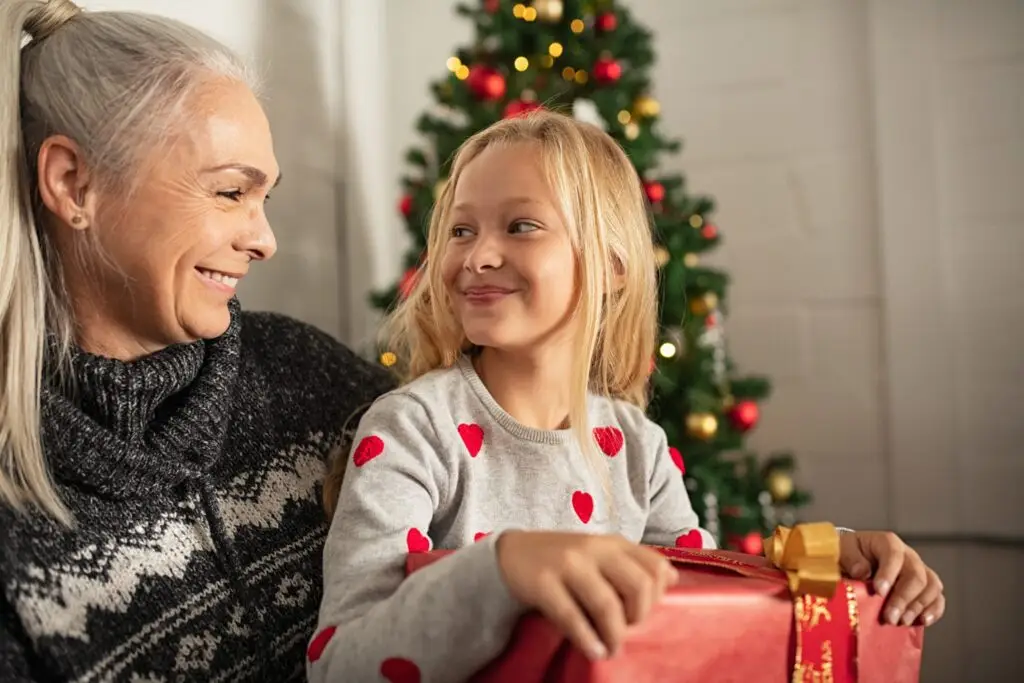 A grandmother and her granddaughter share a joyful moment in front of a decorated Christmas tree. The grandmother dons a patterned sweater while the granddaughter, holding a wrapped gift, wears a gray sweater with red hearts—your guide to the best tech gifts this Christmas shining brightly.