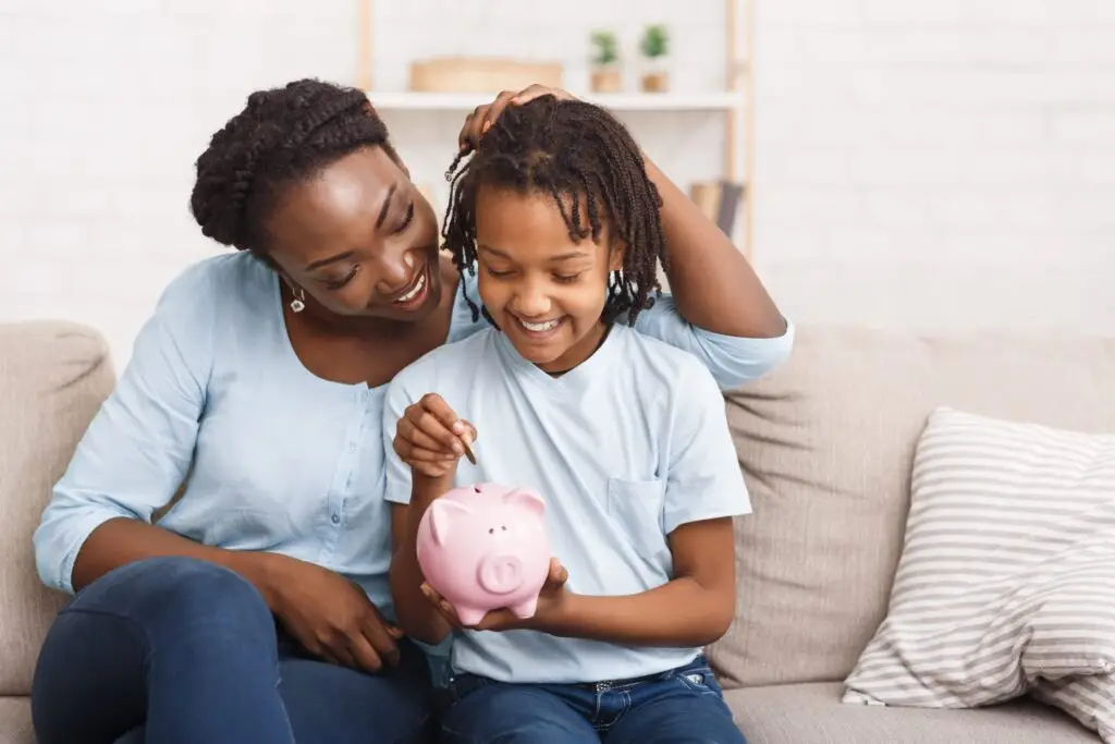 A woman and a child sit on a couch, smiling as the child inserts a coin into a pink piggy bank. Both wearing light blue shirts, they embrace this moment of teaching your kids about money. The background features a white brick wall and potted plants on a shelf.