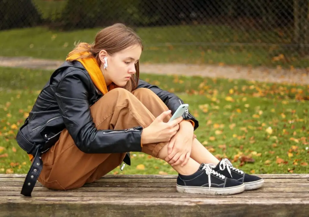 A young person, possibly exploring the safety of messaging apps, sits on a wooden bench outdoors. They wear earbuds, a black leather jacket, and brown pants while intently looking at their phone. Autumn leaves are scattered on the grass in the background.