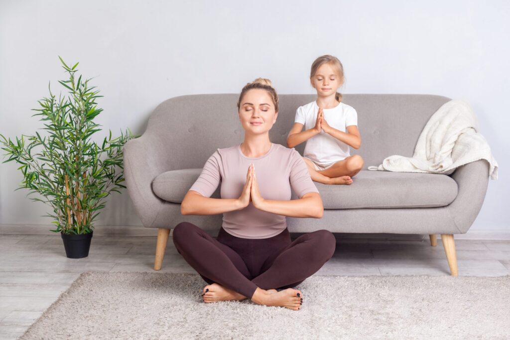 A woman sits cross-legged on the floor with her hands in a prayer position, eyes closed, embracing mental health maintenance. Behind her, a child mirrors her posture on a gray sofa. A potted plant is to the left, and a light rug covers the floor, creating a serene space reminiscent of an app for tranquility.