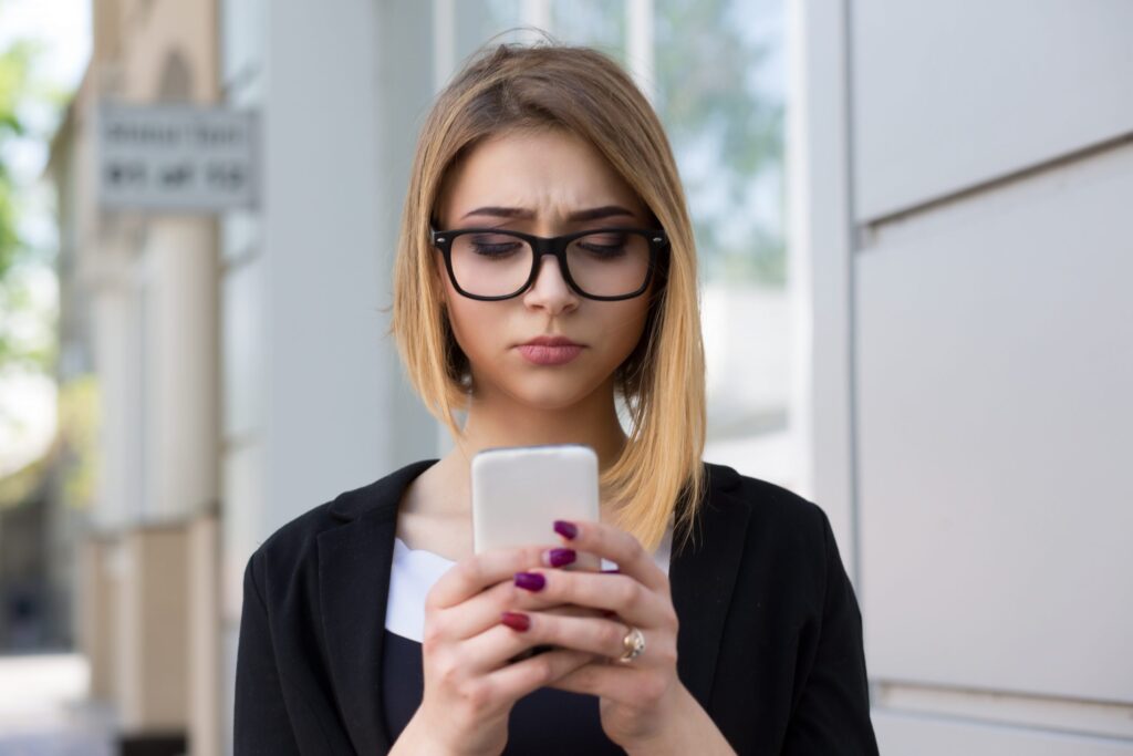 A woman with glasses looks intently at her smartphone while standing outdoors, perhaps pondering, "Is Omegle safe for kids?" She has shoulder-length blonde hair, wearing a black blazer against a backdrop of a blurred street scene with a building.