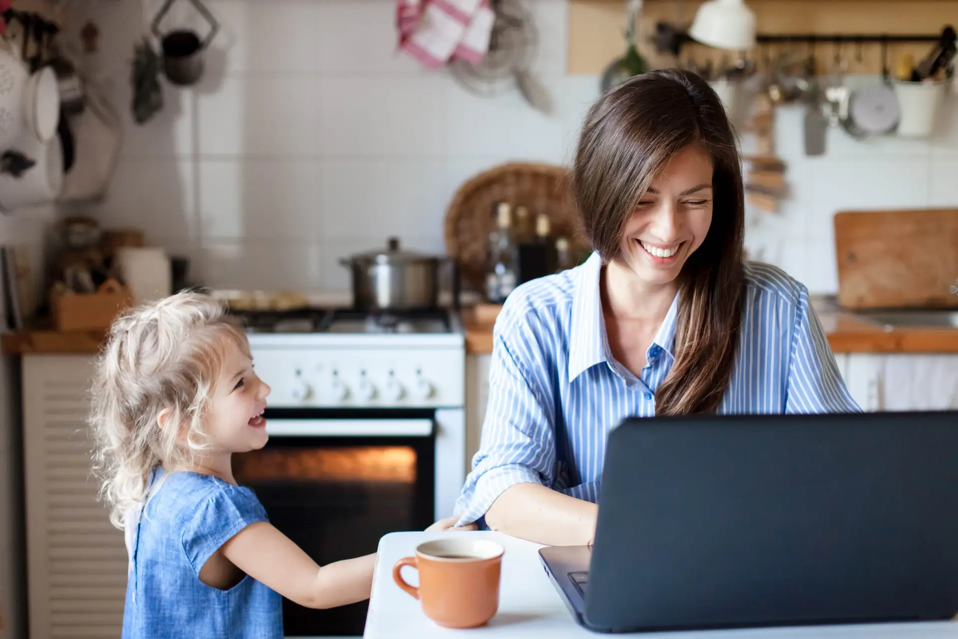 A woman with long brown hair smiles while balancing work and life at the kitchen table, her laptop open. A young girl with curly hair in a blue dress stands smiling beside her. A coffee mug rests on the table, and the cozy kitchen is visible in the background.