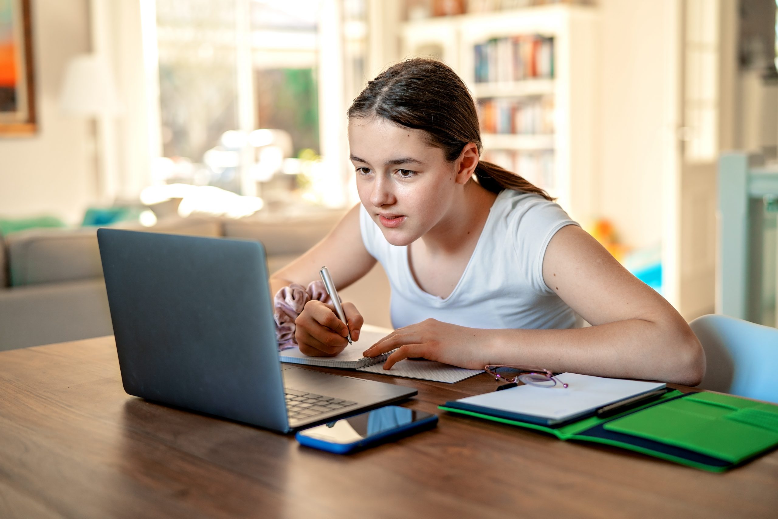 A young person is focused on taking notes while looking at a laptop screen, possibly exploring how to prevent cheating in the digital age. They sit at a table in a well-lit room with bookshelves in the background, complemented by a smartphone and glasses resting on the table.