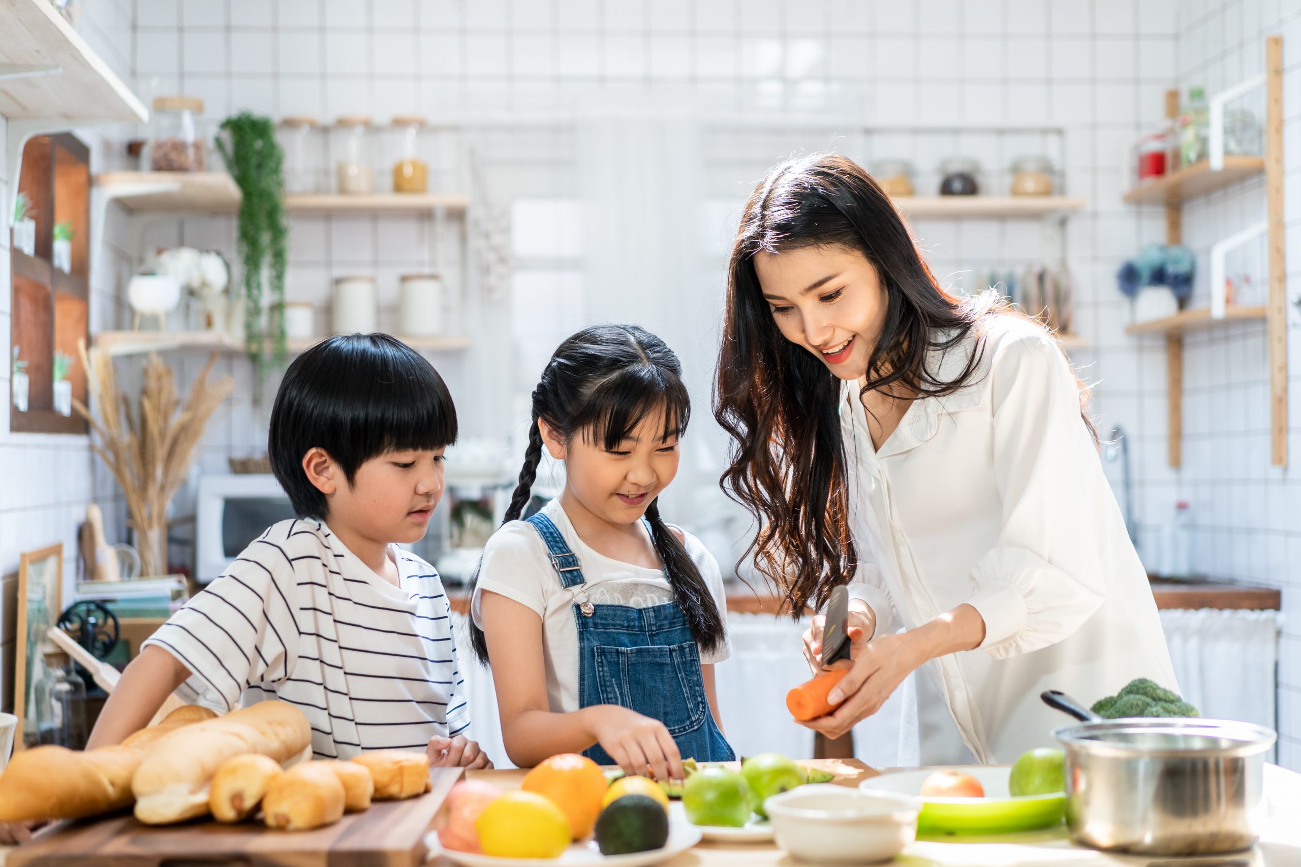 In a bright kitchen, a woman and two children prepare a meal together. As the woman slices a carrot, various fruits and vegetables add color to the counter. Enthralled in this "Let's Cook!" moment, the children smile, embodying simple tips for cooking with kids of all ages. Rolls of bread peek out invitingly.