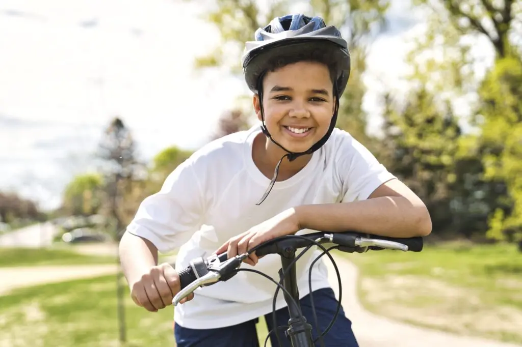 A young boy is smiling beneath his helmet, embracing the joy of trying new things as he rides a bicycle on a sunny day. The trees and grass in the background add to this perfect moment of adventure and discovery.