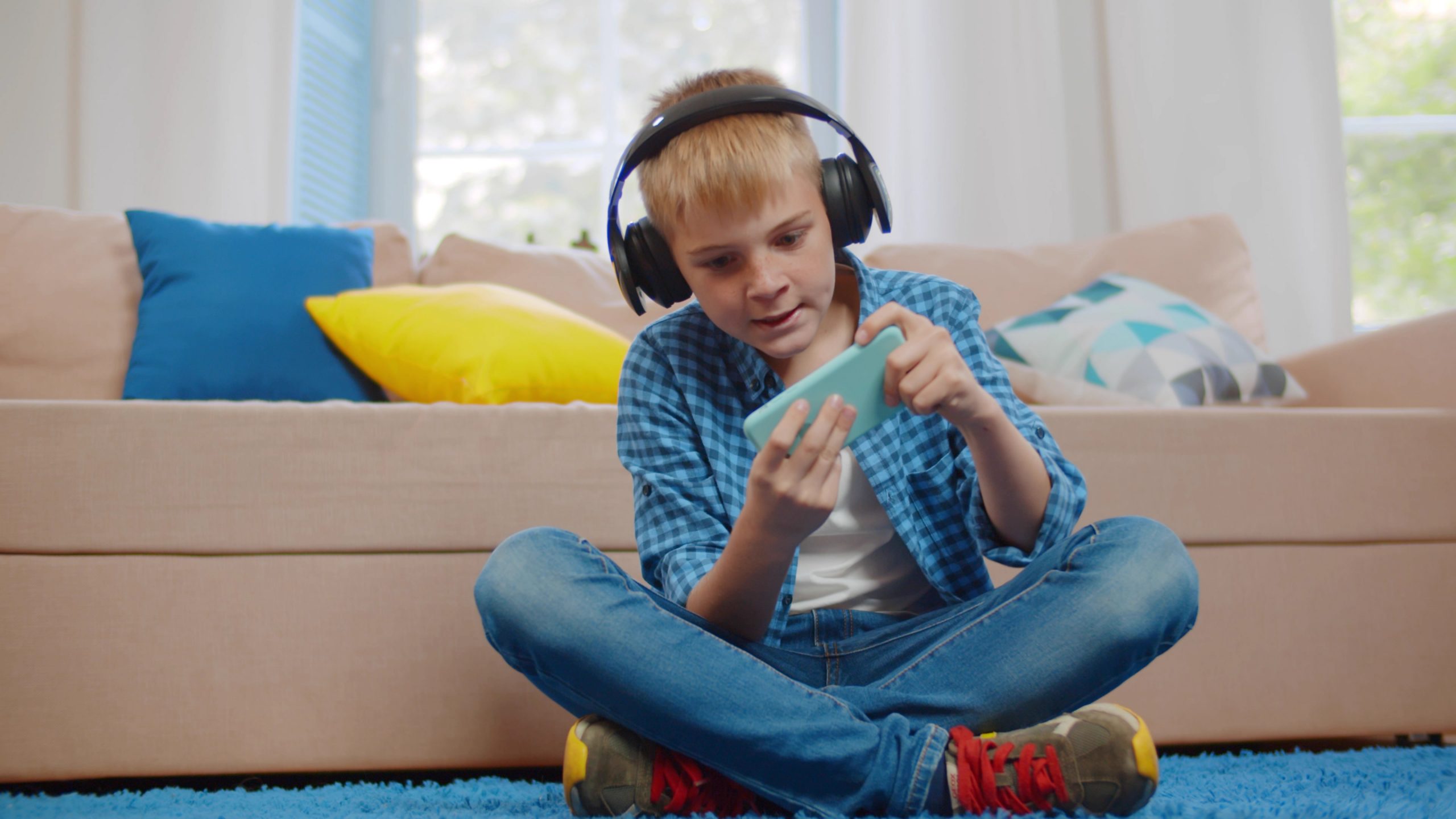 A young boy with headphones sits cross-legged on a blue carpet, playing a game on his handheld device. Clad in a blue plaid shirt and jeans, he's immersed in the moment. Behind him, a beige sofa with colorful cushions prompts the question: should you go for those enticing in-game purchases?