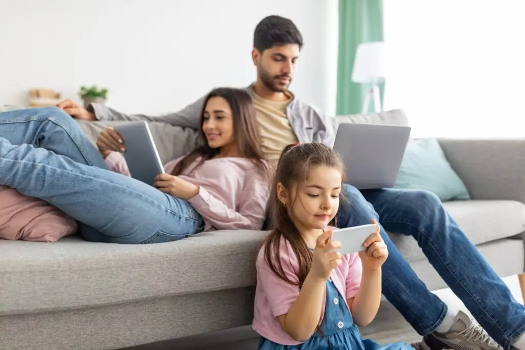 In a casually modern home, a family of three unwinds. A man and woman sit on the couch immersed in their screens, while a young girl on the floor is absorbed in her smartphone. It's a serene scene, yet one might ponder: What is phubbing and why is it bad for relationships?