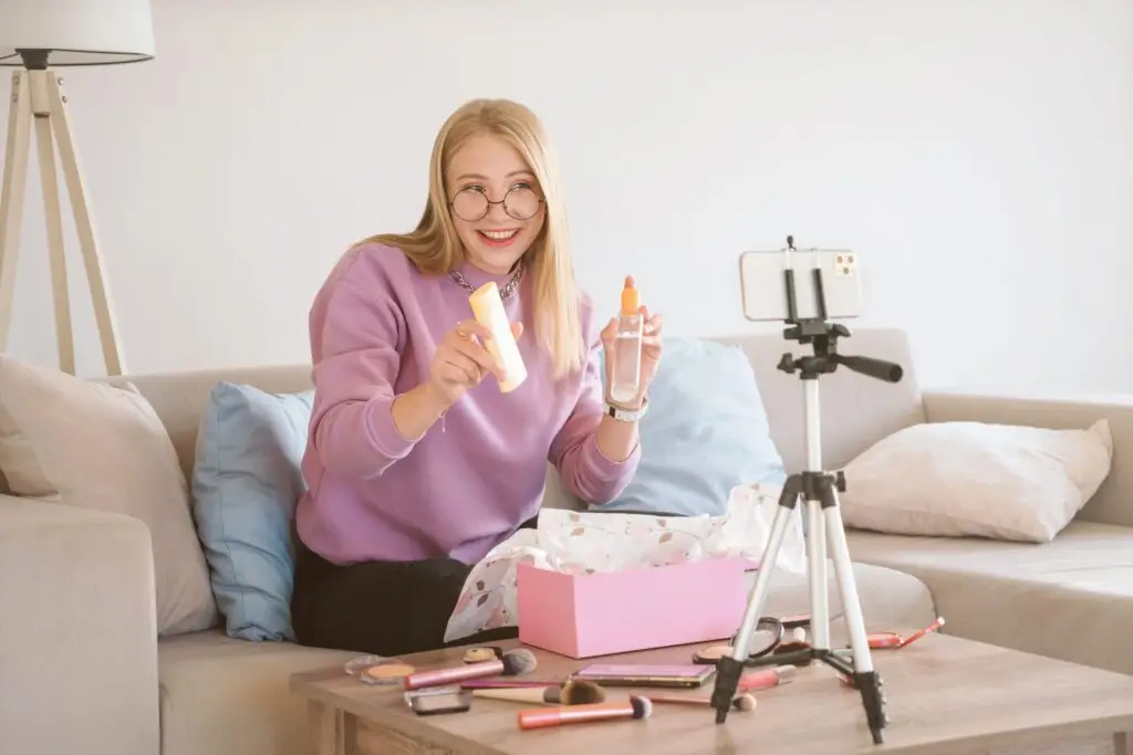 A woman with long blonde hair and glasses sits on a couch, smiling at a smartphone on a tripod. She holds up two cosmetics, surrounded by makeup items on the table. Wearing a purple sweater, she's filming for her #Trending YouTube unboxing video or beauty livestream.