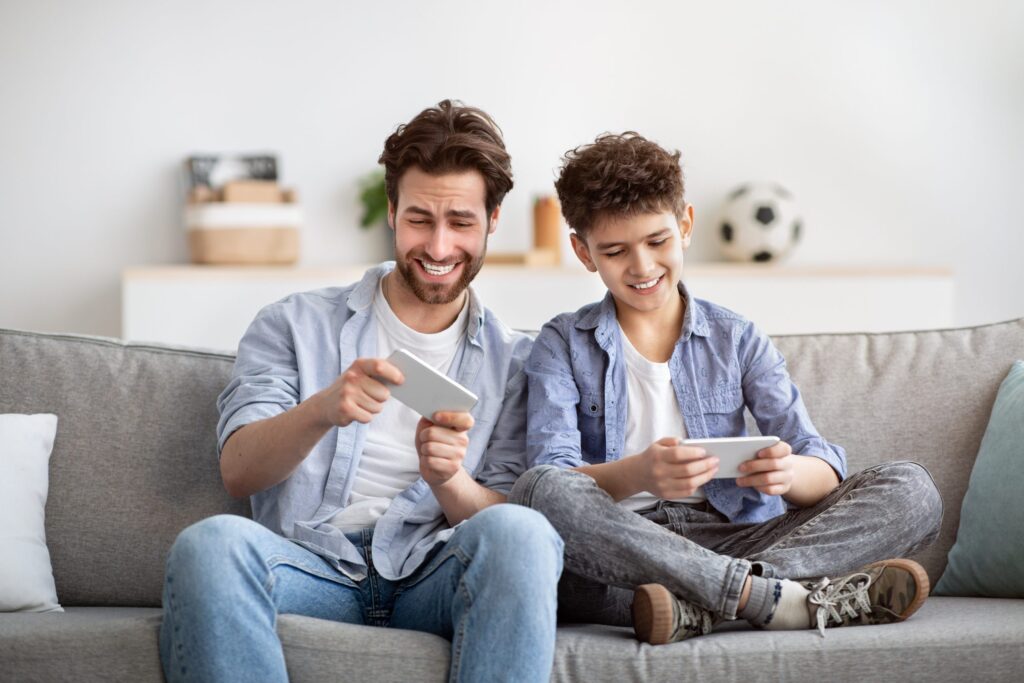 A man and a boy are sitting on a couch, smiling as they explore how Troomi’s smartphone games work. Casually dressed, they enjoy their game time against a soft-focus backdrop of shelves and a soccer ball.