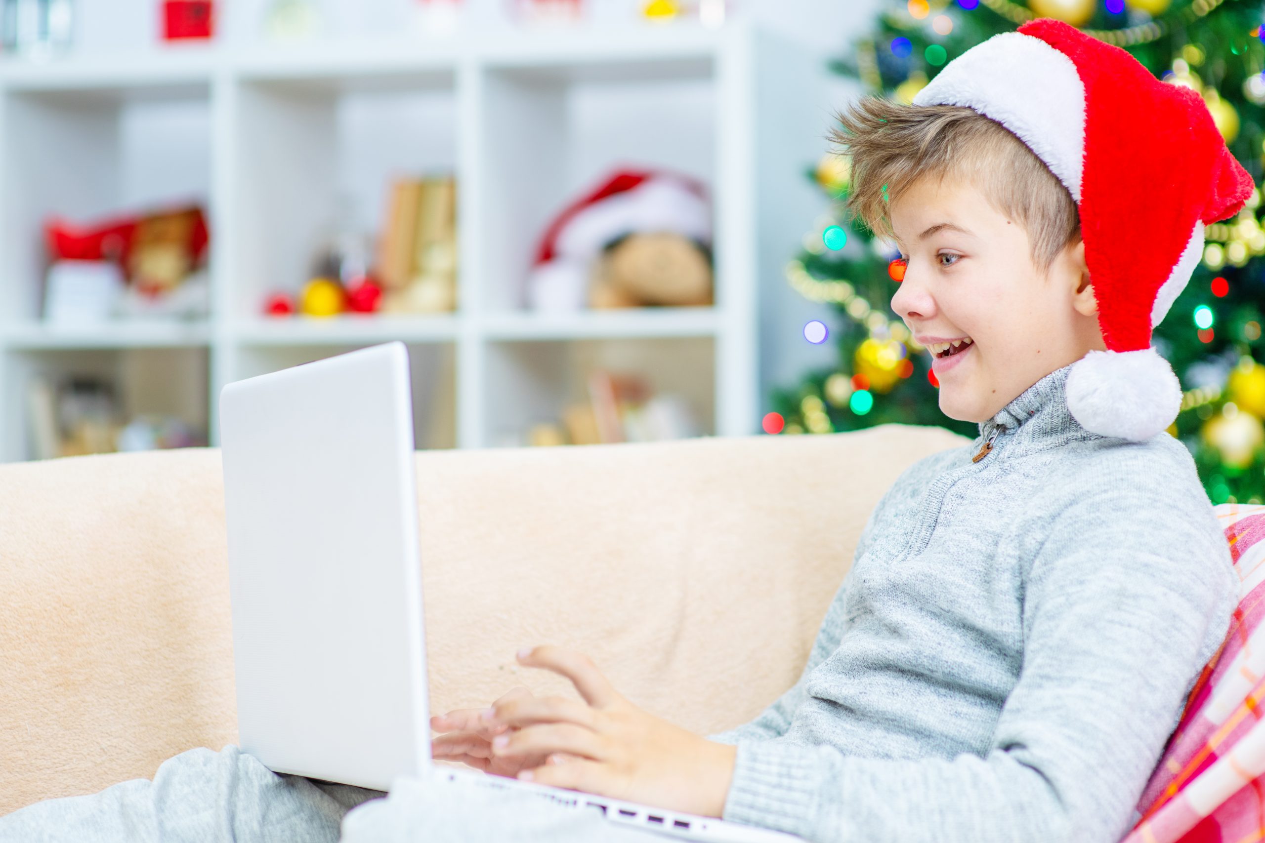 A smiling child wearing a Santa hat and a grey sweater sits on a couch using a laptop, embraced by the cozy holiday spirit. A Christmas tree with colorful decorations twinkles in the background, creating a festive atmosphere while subtly reminding us of tech use during the holidays.