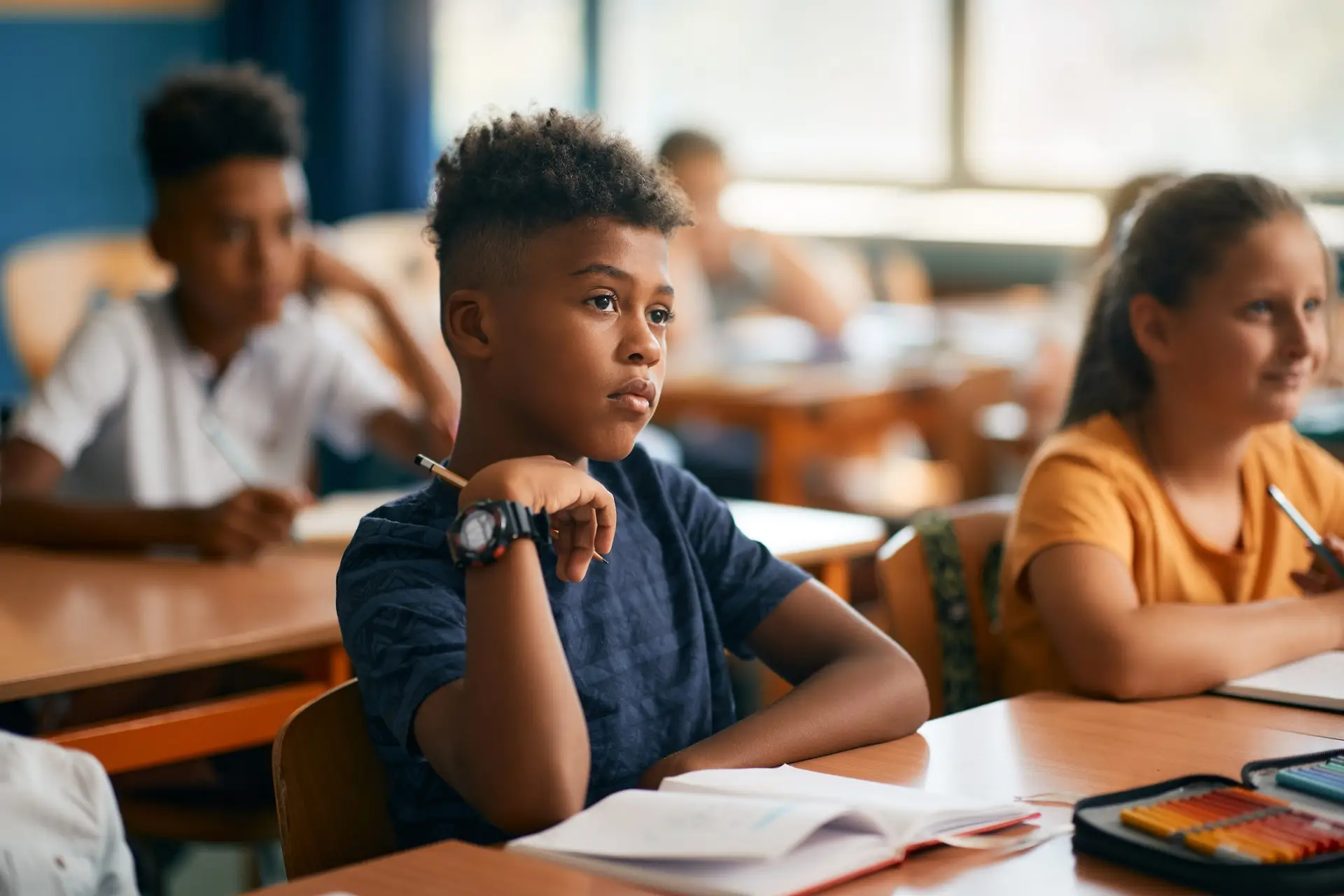 A young student, powered by concentration tips to help your kids stay focused, sits attentively at a classroom desk, with a notebook open in front of him. Other students are seated at desks in the background, partially blurred. The well-lit classroom features large windows.