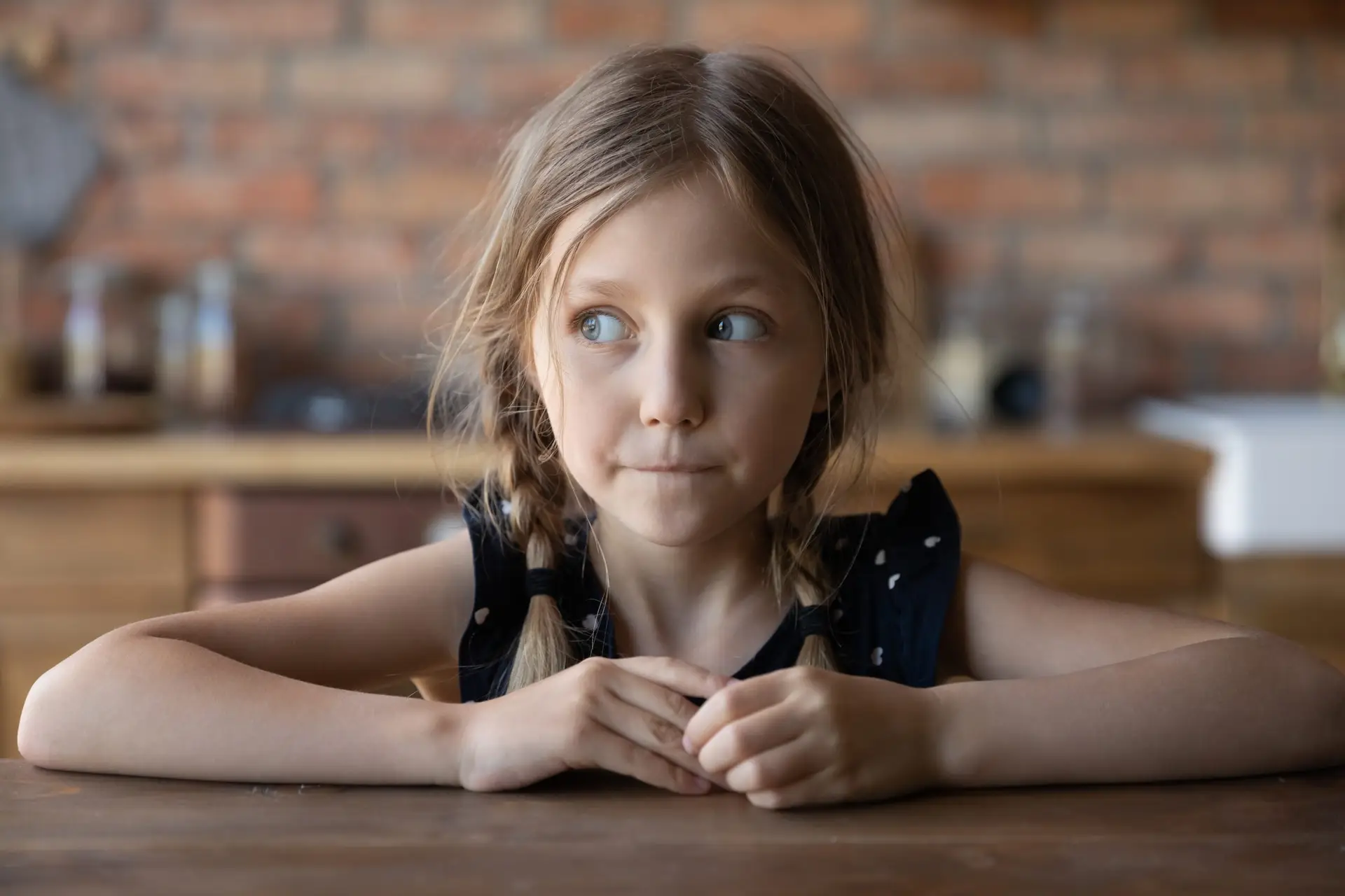 A young girl with braided hair sits at a wooden table, looking thoughtfully to the side, perhaps pondering recent trends like The Tide Pod Challenge. She is wearing a black sleeveless dress. The background features a brick wall and a blurred kitchen setting.