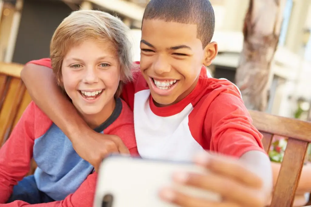 Two boys are sitting on a wooden bench, smiling as they take a selfie together with their cell phones. One boy has his arm around the other. They look happy and are wearing casual t-shirts, showing how technology can capture joyful moments in kids' lives. The background is slightly blurred.