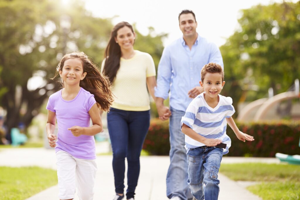 A happy family of four enjoys a tech-free day in the park. Two children, a girl and a boy, run ahead, smiling wide. Behind them, their parents hold hands and savor the moment. The sun shines down as trees sway gently in the background, offering simple tips to decrease tech consumption naturally.