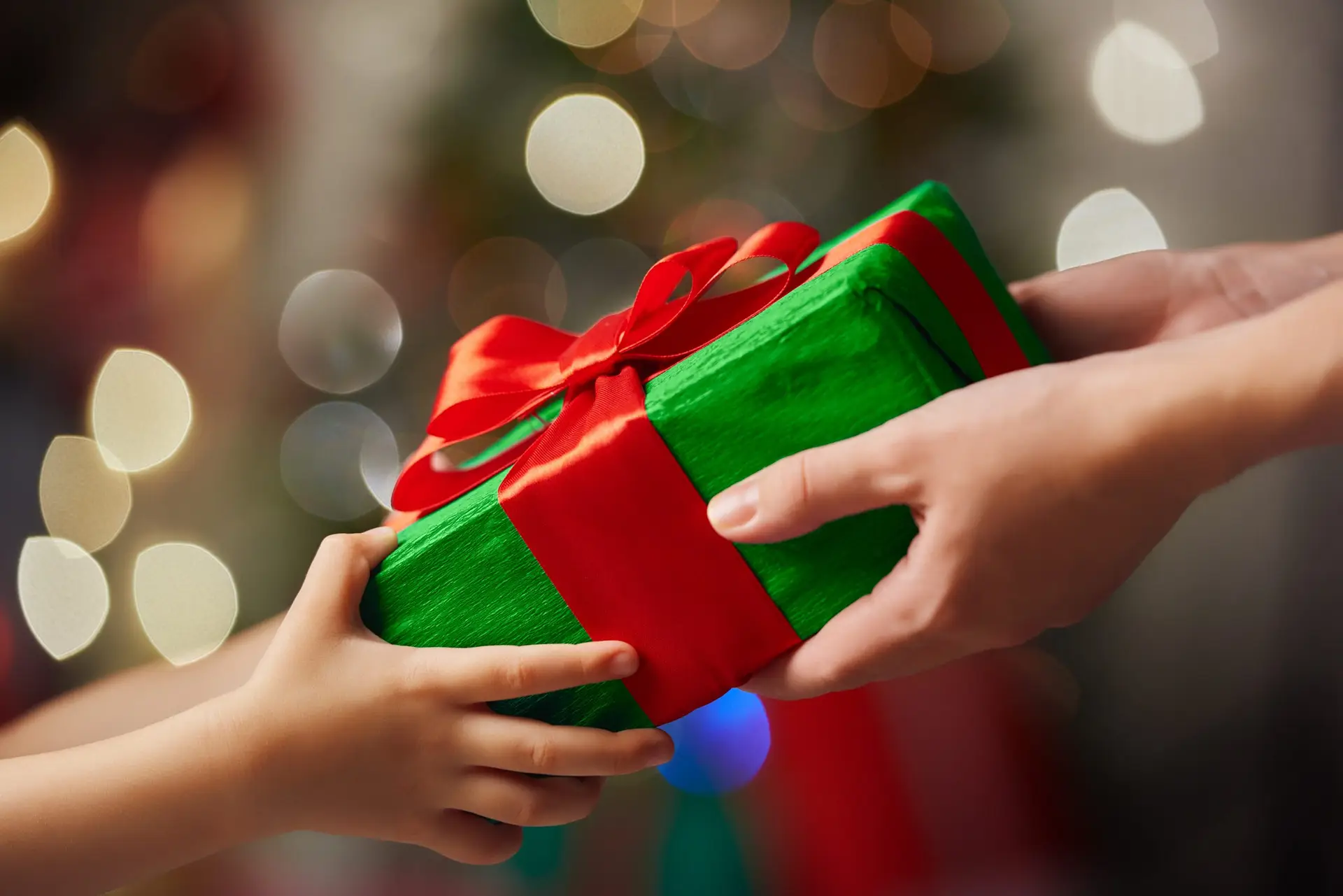 Two hands exchanging a wrapped gift, teaching kids the joy of giving at Christmas. The present, adorned in green wrapping paper and tied with a red ribbon, sits at the heart of this warm tradition. Soft-focus lights in the background create a festive atmosphere.