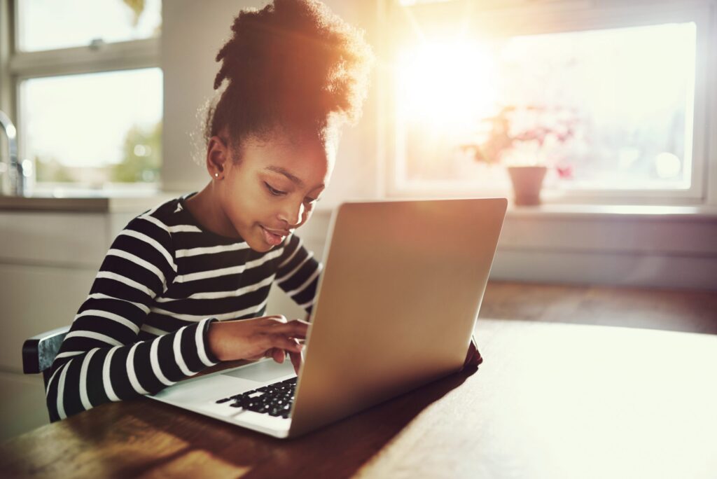 A young girl in a striped shirt sits at a table, engrossed in "Don’t Pour Juice on the Laptop: Computer Basics for Kids" as she uses her laptop. Sunlight streams through the window, illuminating her and creating a warm ambiance.