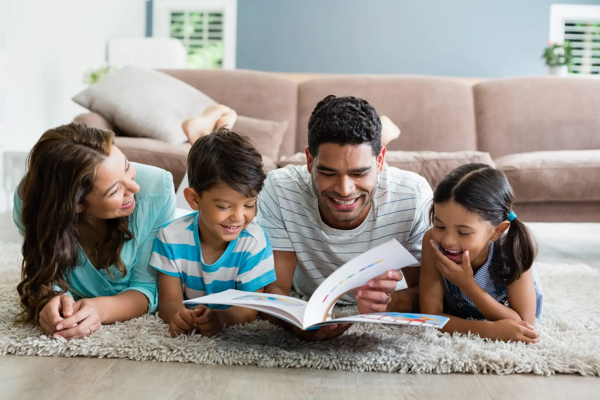 A family of four, basking in the warmth of their cozy living room, lies on the carpeted floor, smiling and reading "10 Hobbies You and Your Kids Can Start at Home This Winter." The parents and two young children are happily engaged in discovering new activities together.