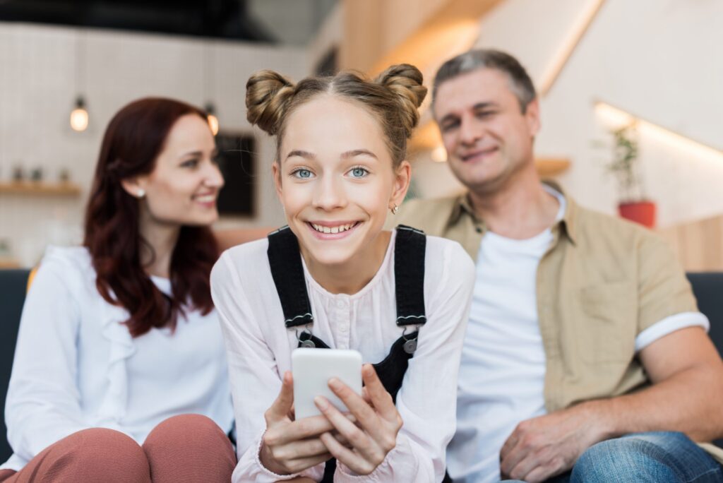 A young girl with hair in two buns smiles while holding a smartphone, embodying the innocence that we strive to protect. A woman and a man sit in the background, smiling warmly in their cozy, modern living room, fostering an environment where kids can safely explore technology.