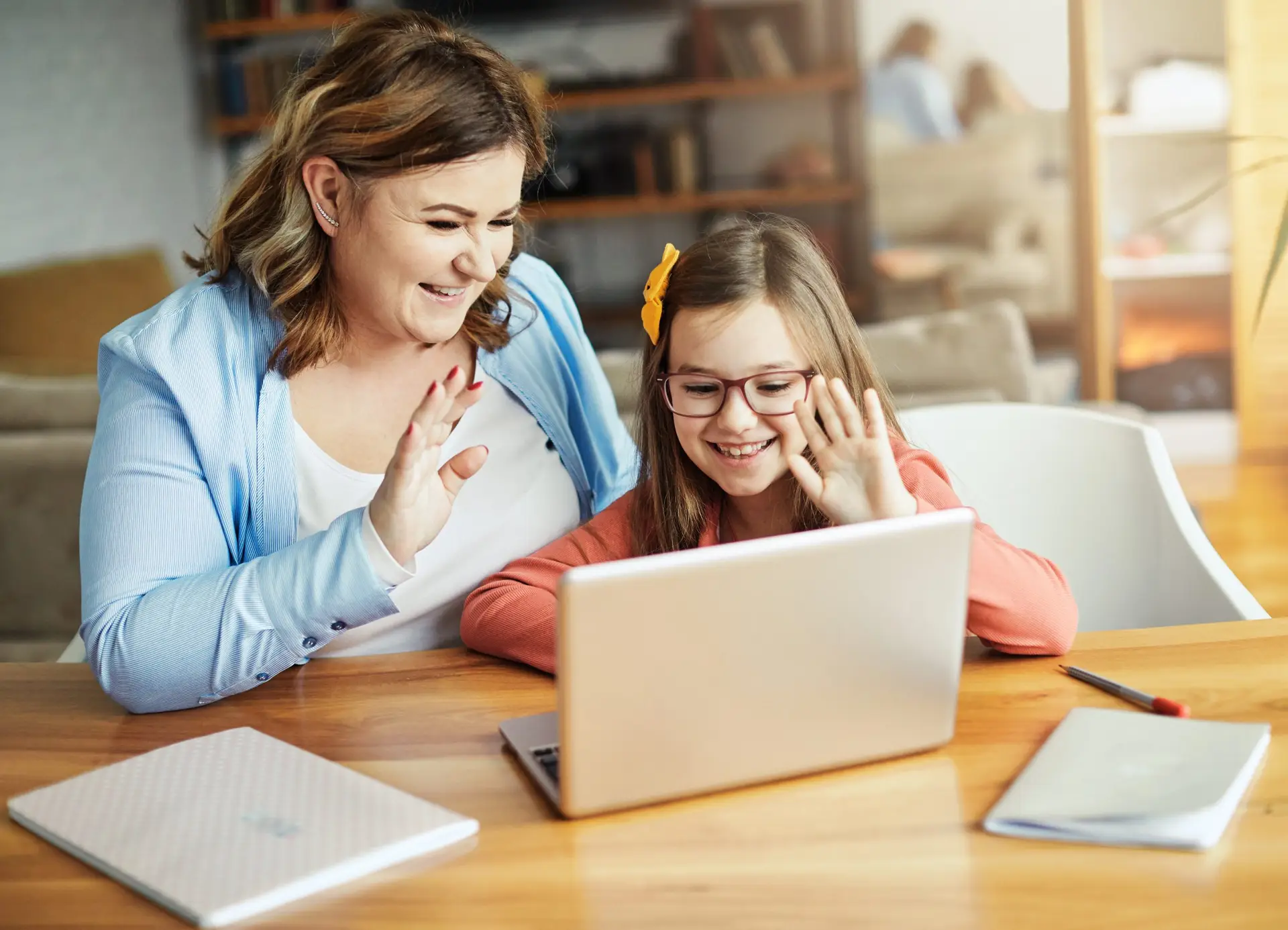 A woman and a young girl are sitting at a wooden table, smiling and waving at a laptop screen, learning how to digitally talk with your kid’s teacher. The woman is in a blue shirt, while the girl with glasses and a bow in her hair has a notebook and pen ready nearby.