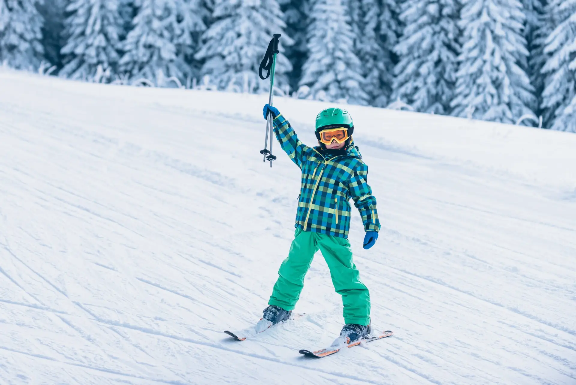 A child in green ski gear and a blue checkered jacket holds up a ski pole while skiing down a snowy slope, showcasing one of the 5 ways spending winter outdoors benefits kids’ wellbeing. Snow-covered trees provide a serene backdrop to this joyful embrace of nature.