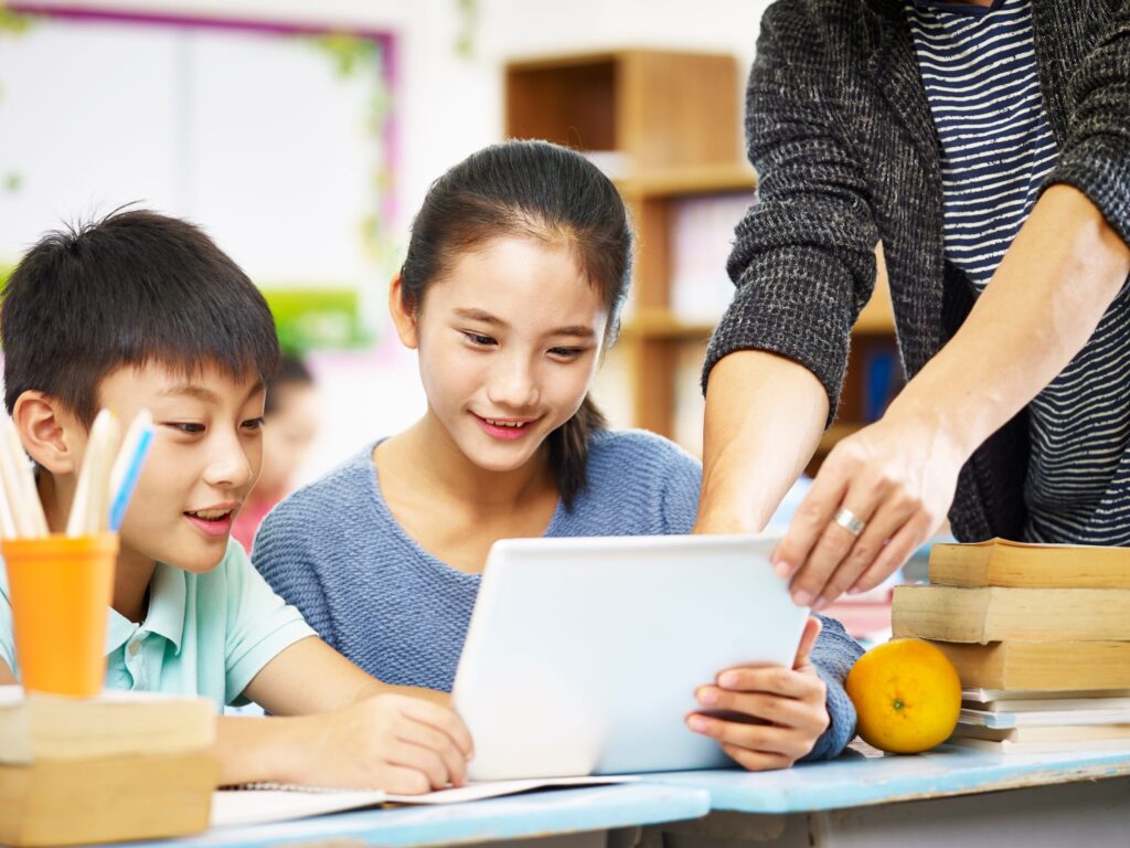 Two children in a classroom are exploring educational apps on a tablet screen held by their teacher. The boy wears a light blue shirt, and the girl is in a blue sweater. Books and an orange are on the table, while shelves and a colorful bulletin board add vibrant background details.