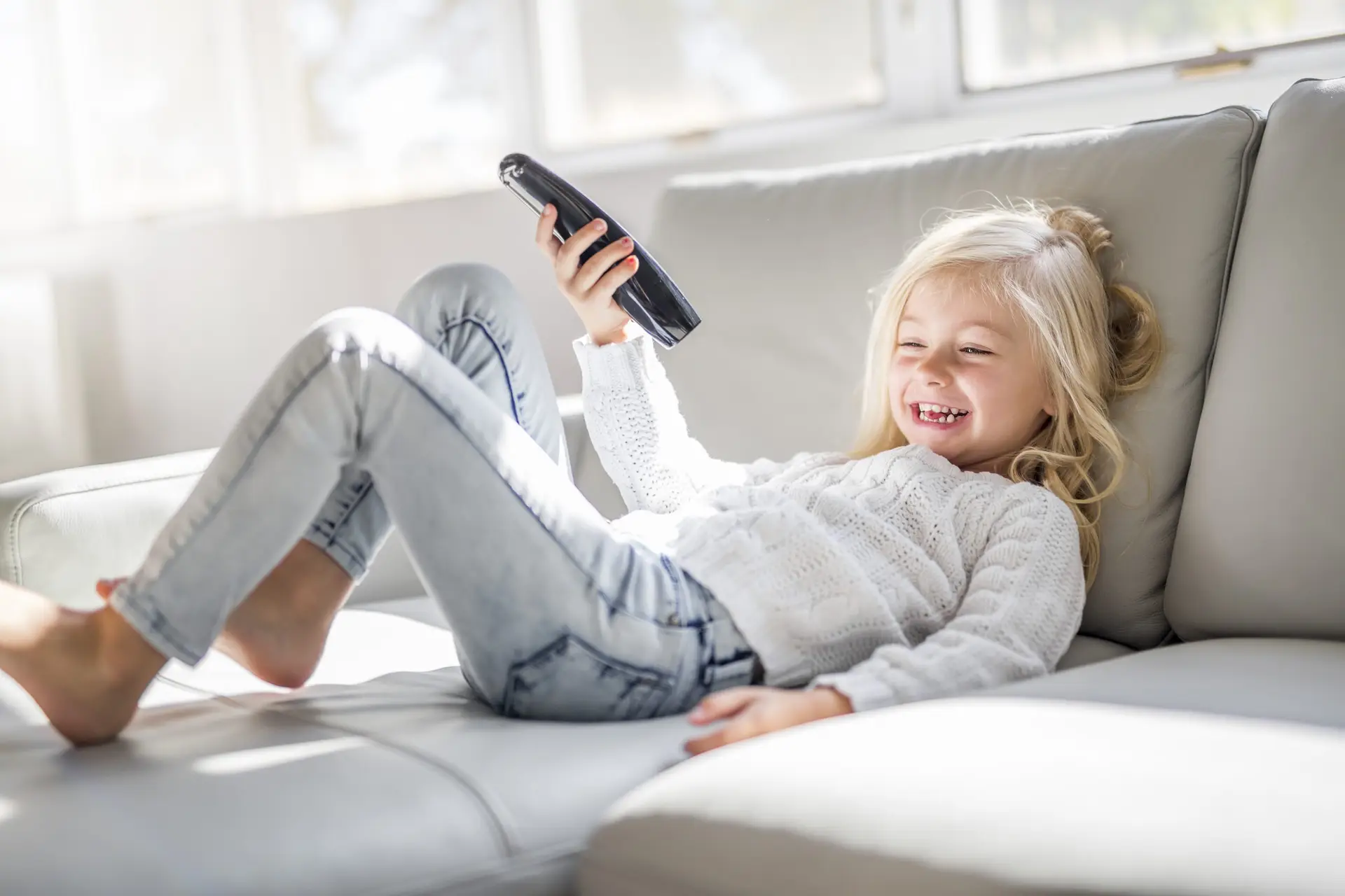 A young girl with blonde hair relaxes on a light-colored sofa, holding a remote control and smiling, as she tunes in to the news. She wears a white sweater and light jeans. Sunlight streams in through the windows, offering parents a moment to ponder if watching the news is good for kids.