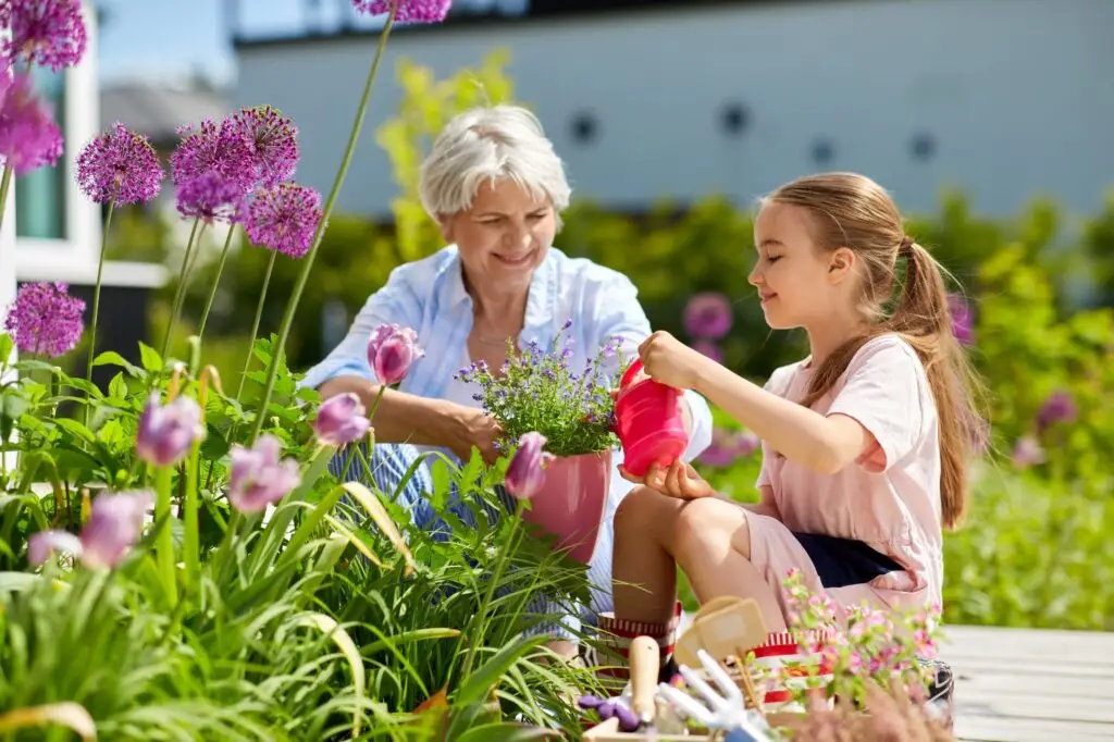 A woman and a young girl enjoy gardening together, embodying the six reasons to teach your kids how to garden and love it. The woman holds a potted plant while the girl waters it with a small red watering can, surrounded by vibrant flowers and greenery on this sunny day outdoors.