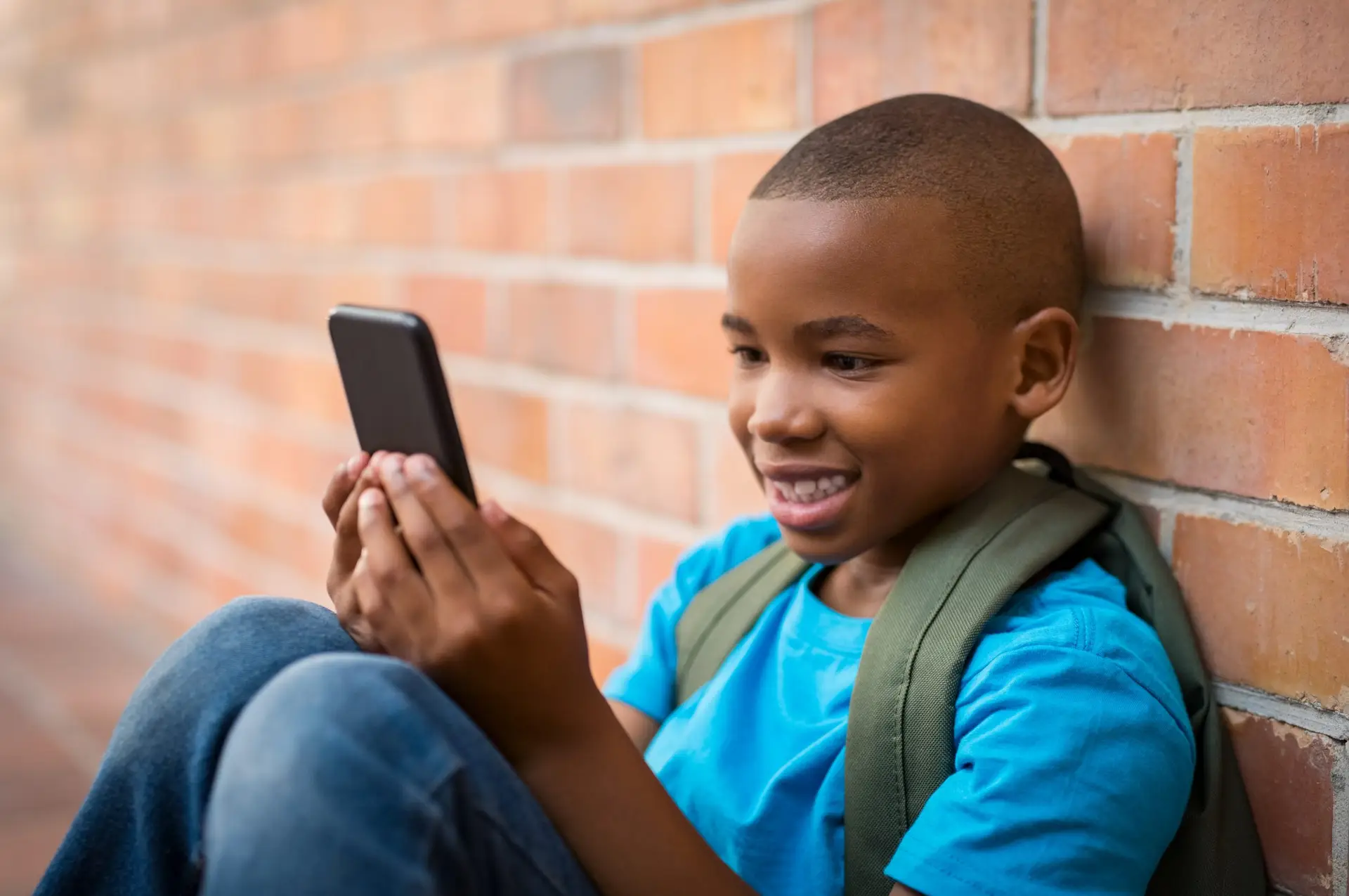 A smiling boy with a short haircut, wearing a blue shirt and green backpack, sits against a brick wall, engrossed in his smartphone. With Troomi Phones' careful design ensuring no pornography ever reaches young eyes, parents can trust this safe tech companion.