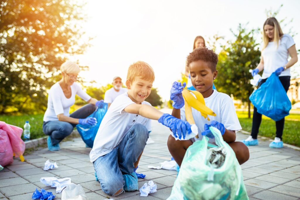 Children and adults in a park are wearing gloves and cleaning up litter, exemplifying how you can help your child become civically engaged at any age. Two boys focus on placing trash into a bag, while others gather waste in the sunny, tree-lined scene.