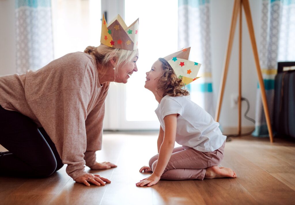 A woman and a young girl kneel on the wooden floor, wearing colorful paper crowns. They lean toward each other, smiling in a bright room with curtains and a standing lamp in the background—capturing their joyful moment during the weekly family fun night to beat the winter blahs.