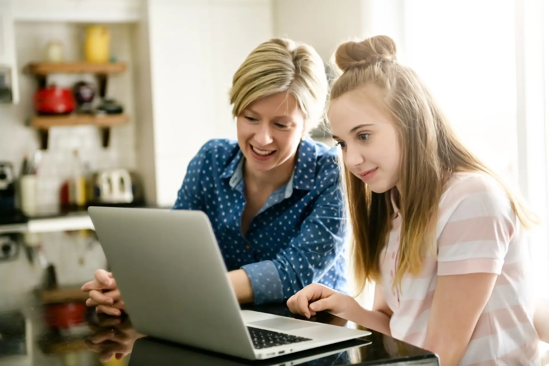 A woman with short blonde hair in a blue polka dot shirt and a girl with long hair in a top knot sit at the kitchen counter, focused on their laptop. They seem absorbed, possibly exploring online topics like "What is a VPN?: Virtual Private Networks Explained.