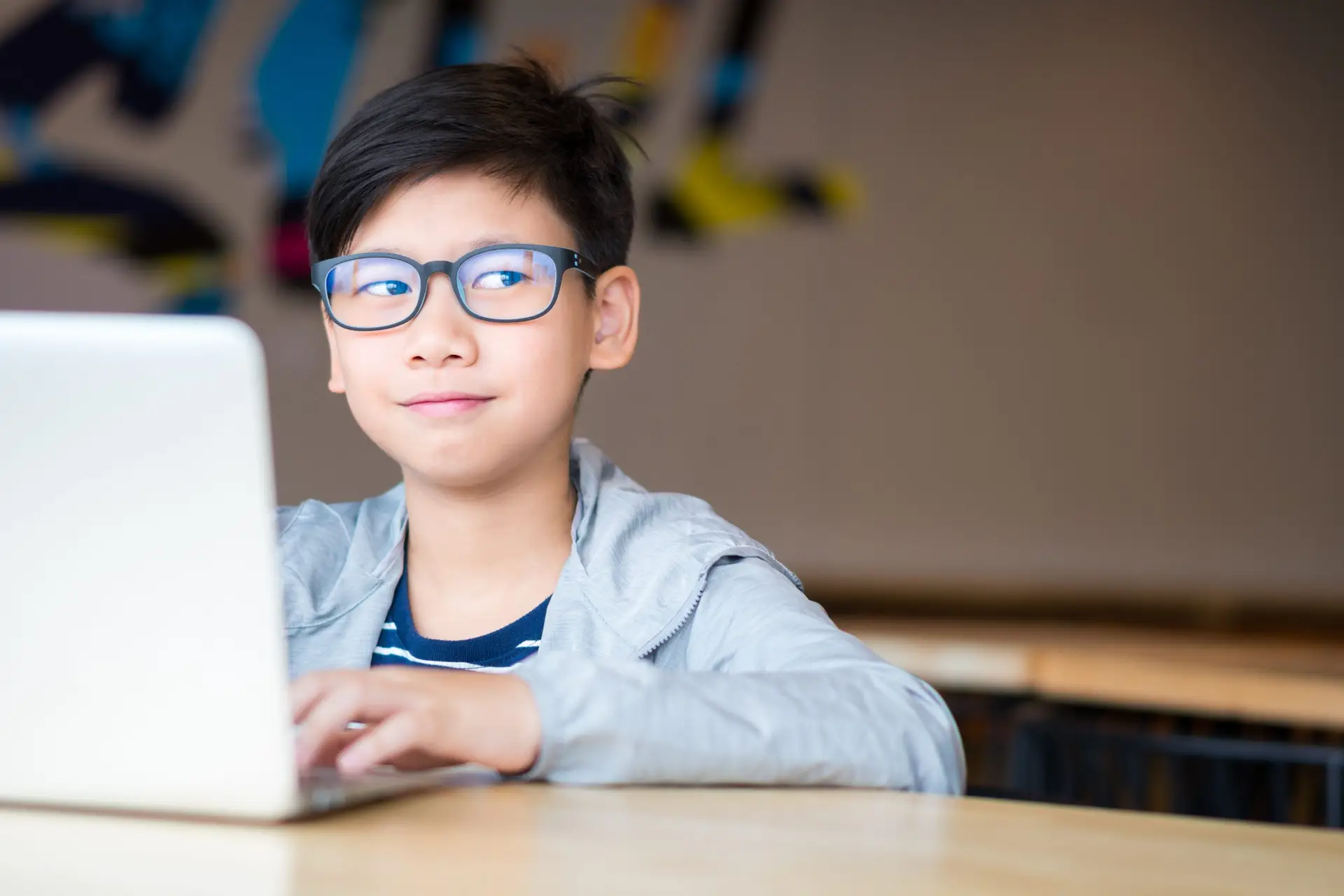 A young boy wearing blue light glasses sits at a wooden table using a laptop. He is smiling slightly and looking to the side. The background is blurred, with colorful abstract shapes on a wall, proving these glasses are more than just a trend.