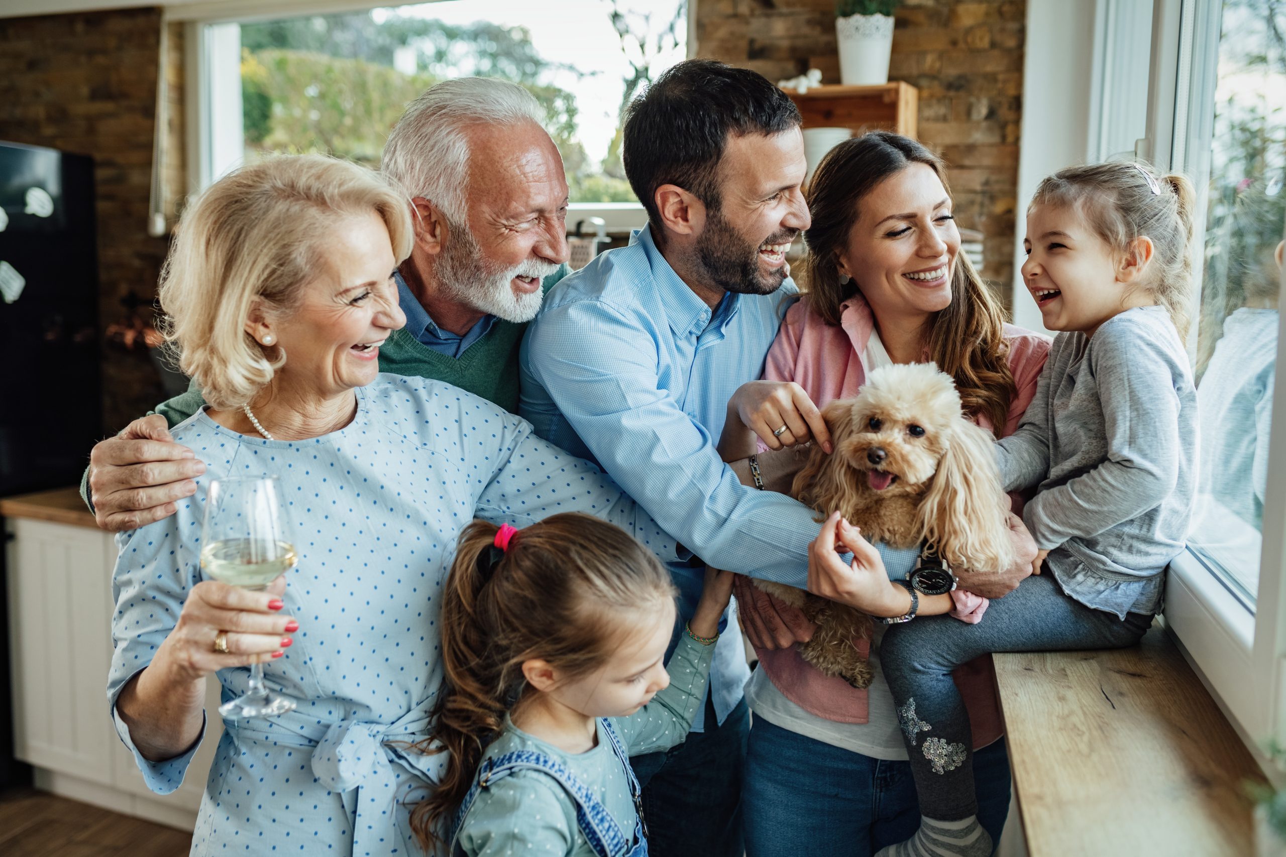 A joyful family gathers in a kitchen, embodying the essence of how (and why!) to keep in touch with your extended family. An older couple smiles with an adult man and woman, who hold a small dog and a laughing child. Another child looks on, reveling in the happy, relaxed atmosphere.