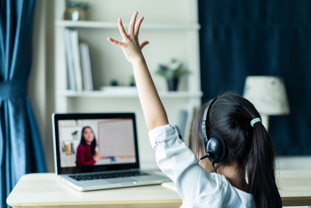 A young girl wearing headphones sits at a desk, raising her hand while attending an online class on her laptop. A teacher is visible on the screen, standing near a whiteboard, sharing tips on how to change your Zoom background like a pro. The room has bookshelves and a lamp in the background.