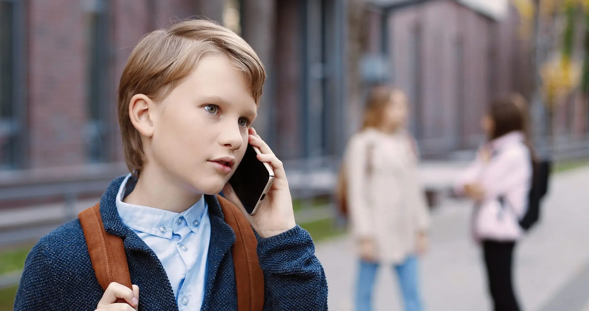 A young boy with light brown hair stands outside, a smartphone pressed to his ear. Wearing a blue sweater and a brown backpack, he stays connected and secure. In the blurred background, two other children chat—thanks to Troomi's safety features keeping kids safe from strangers.