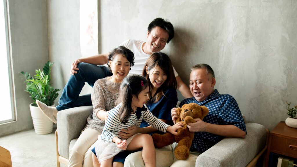 A joyful family sits together on a couch in a cozy living room, filled with warmth and love. A little girl in a striped shirt holds a teddy bear while smiling adults, including the woman who always shares tips for creating healthy boundaries with your family, surround her. A potted plant is in the background.