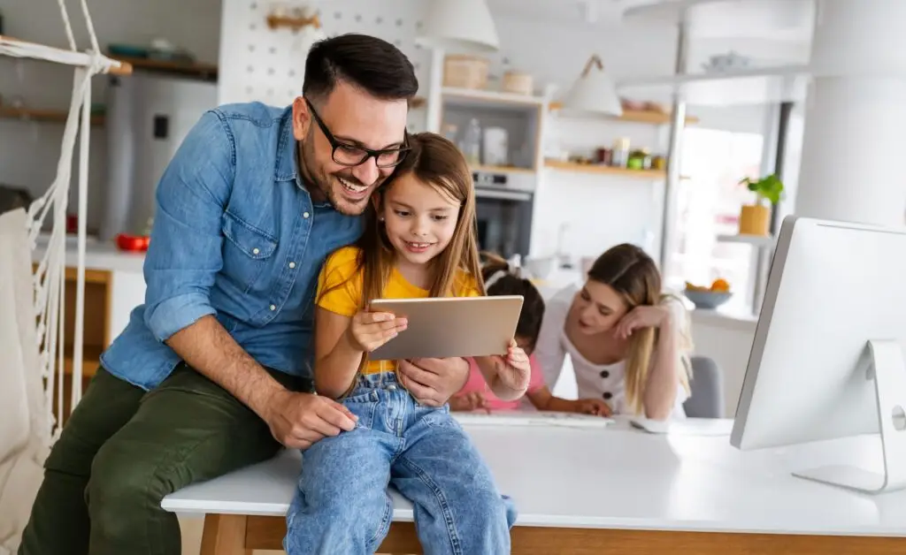 A man and a young girl, embodying the essence of digital citizenship, smile as they explore a tablet at the kitchen counter. In the background, a woman is focused on her laptop. The modern and well-lit kitchen provides an ideal setting for understanding what it means to be part of the digital world.