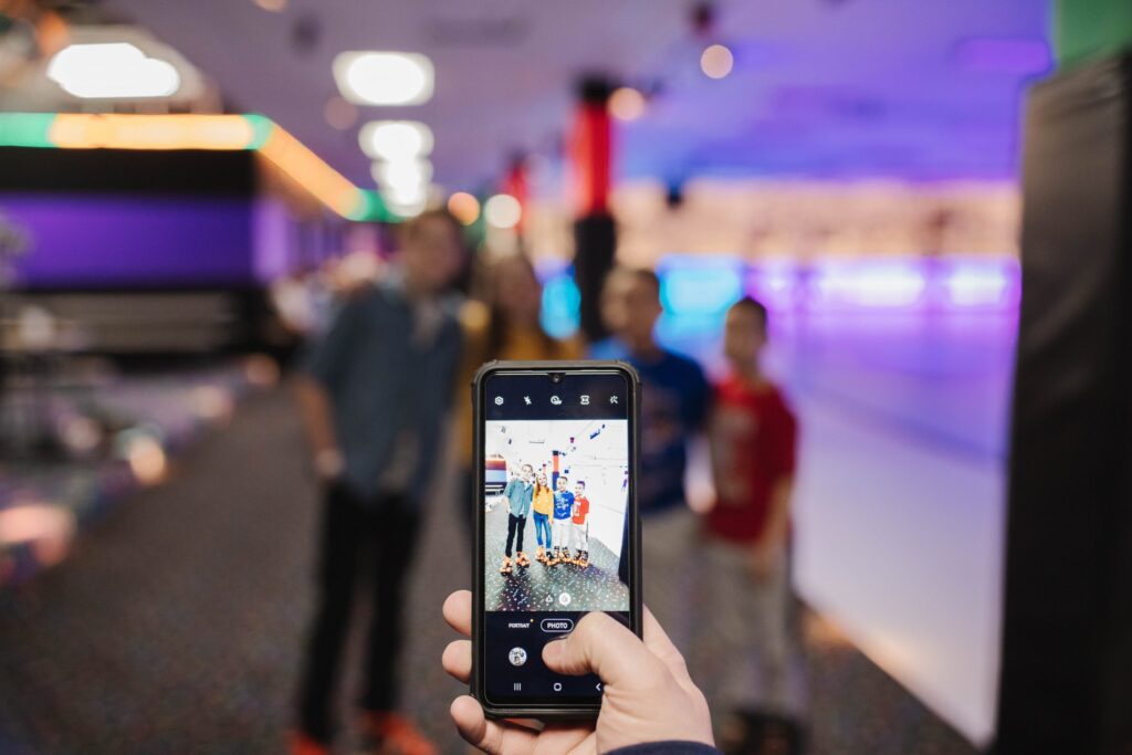 A person holds a smartphone, capturing a blurred photo of three people standing in a colorful indoor space. The background, out of focus with neon lights and a patterned carpet, highlights one of the three reasons your child will love the Troomi Phone Camera.