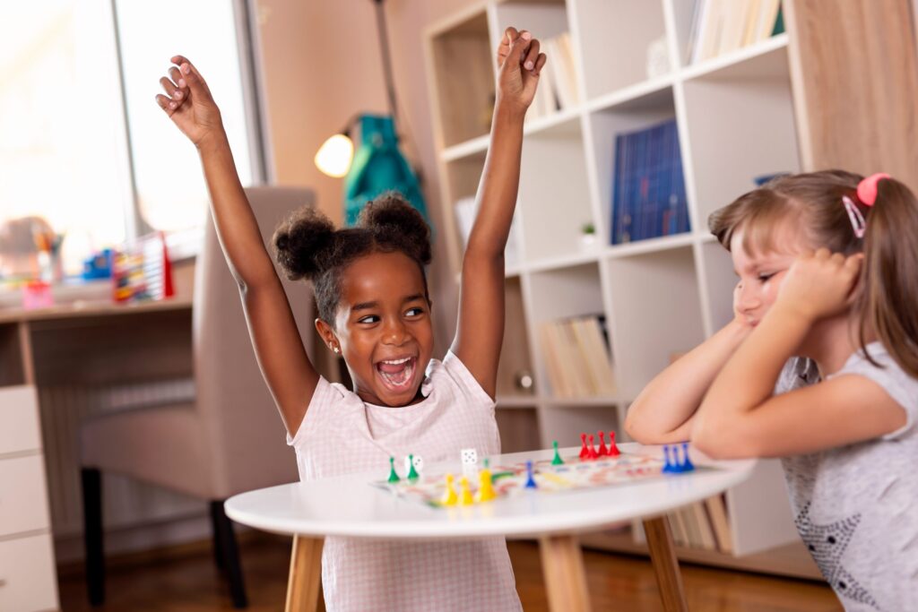 Two young girls are sitting at a small table, absorbed in a board game. The girl on the left is raising her arms, smiling excitedly, while the girl on the right rests her chin on her hands, looking thoughtful. Bookshelves stand in the background—a perfect setting for learning through play and letting kids fail gracefully.