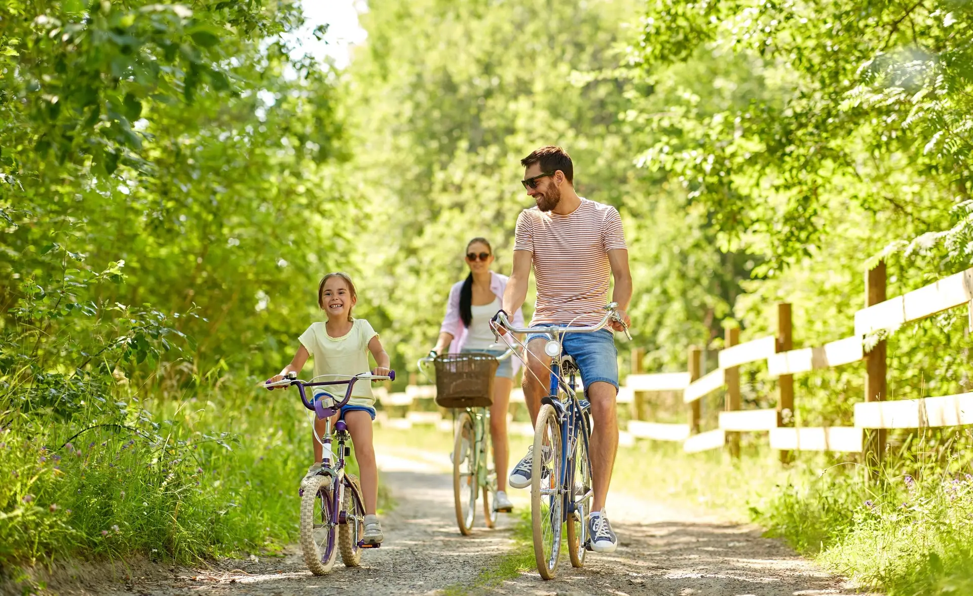 A family of three rides bicycles on a sunny, tree-lined path, showcasing how to keep kids entertained outdoors. The young girl in front smiles while riding, followed by a woman with sunglasses and a wicker basket on her bike, and a man in a striped shirt. The scene is bright and cheerful.