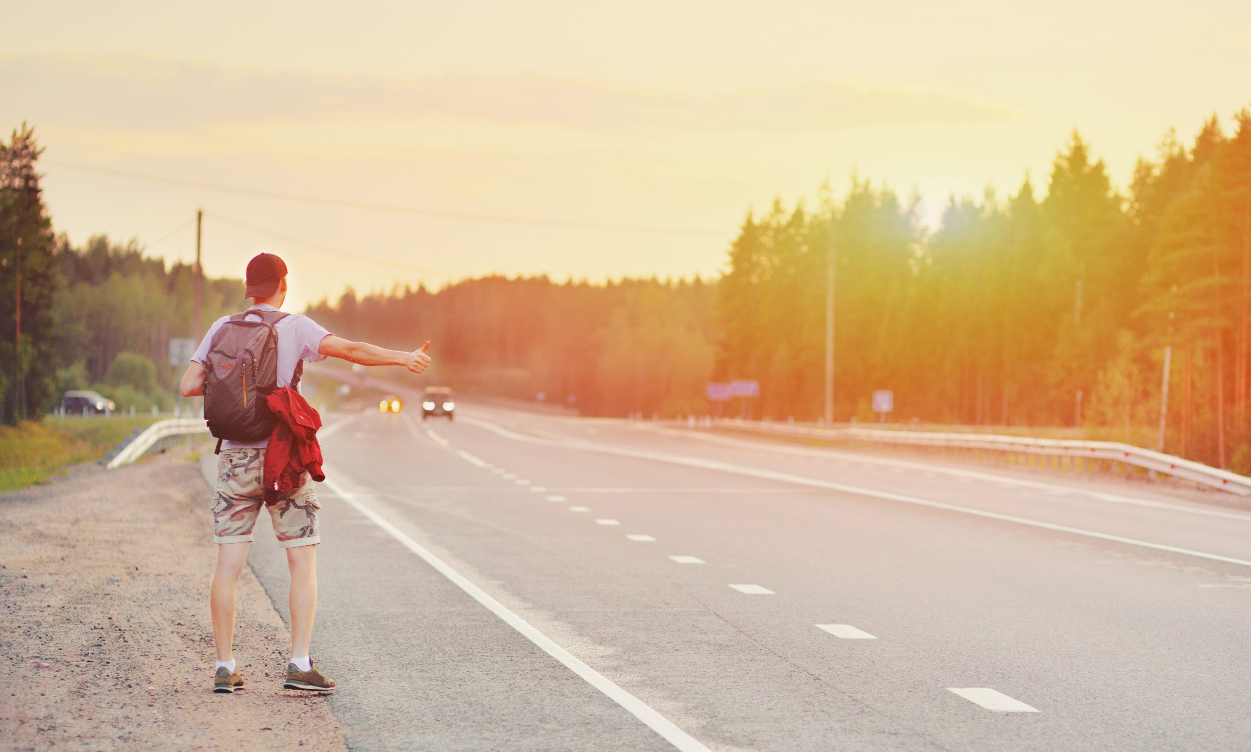 A person wearing a backpack and holding a red jacket stands by the roadside, hitchhiking on a highway. The sun sets, casting a warm glow over the trees and road—a scene far removed from digital threats like drive-by downloads, which travelers should also learn to avoid while exploring.