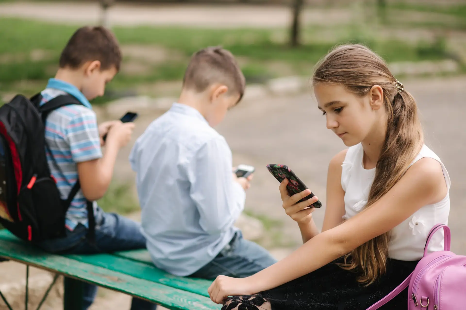 Three children sit on a bench outdoors, engrossed in their smartphones. A girl in a white top and two boys, one in a blue shirt and the other striped, all have backpacks. The green park-like background adds to the scene as they explore why phones are good for kids through educational apps.