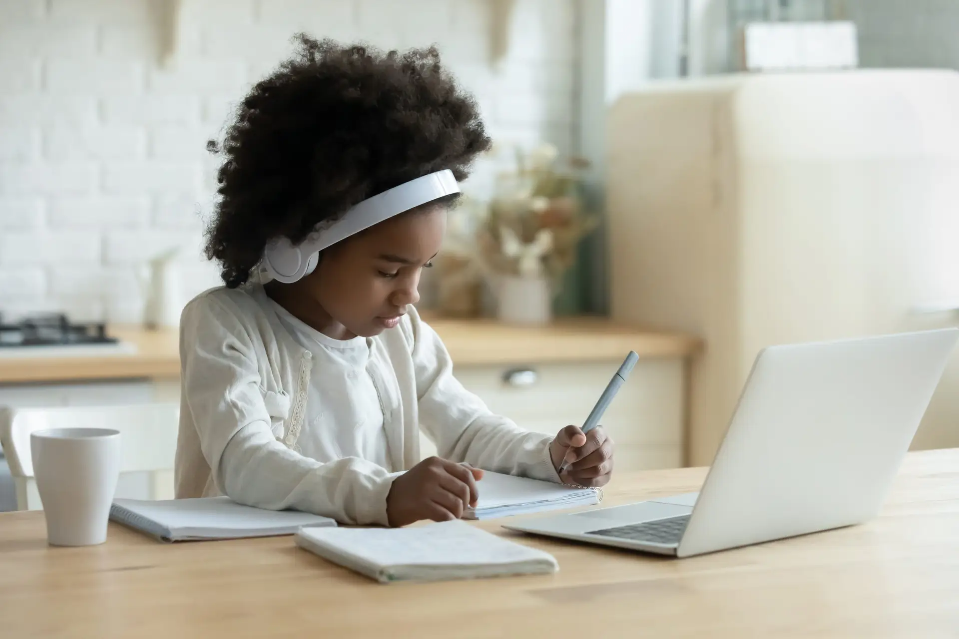 A young child with curly hair sits at a kitchen table, headphones on, deeply focused on writing in a notebook. A laptop lies open alongside a white mug, showcasing how tech has entered the classroom. The bright and casual setting hints at the rise of hybrid schools.