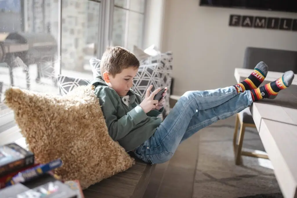 A young boy in a green hoodie and jeans sits on a cushioned bench by a window, focused on a smartphone. With colorful socks and a fuzzy pillow beside him, he relaxes as sunlight streams in. He feels secure knowing Troomi helps stop cyberbullying, creating a cozy atmosphere for screen time.