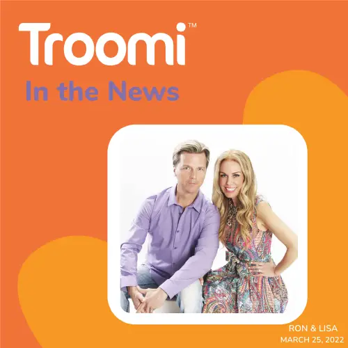 A man and a woman pose together, smiling against a white background. Framed by an orange design with "Troomi In the News: The Gift That Keeps on Giving—Even to Moms" text, the image captures Ron & Lisa with their names and March 25, 2022 date in the bottom corner.