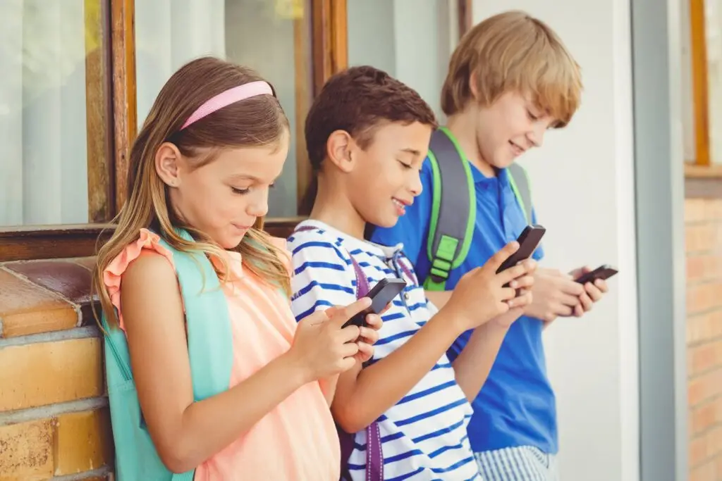 Three children stand side by side, engrossed in their smartphones, possibly exploring the latest killer apps. They wear casual summer clothes and backpacks, with a window and brick wall providing the backdrop to this snapshot of modern life.