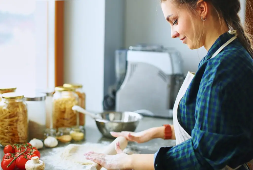 A woman in a plaid shirt and apron rolls out dough on a kitchen counter, mastering life skills for teens. Surrounding her, fresh tomatoes, garlic, and jars of pasta await their turn. A mixing bowl and pasta maker sit ready in the background as natural light streams in from a nearby window.