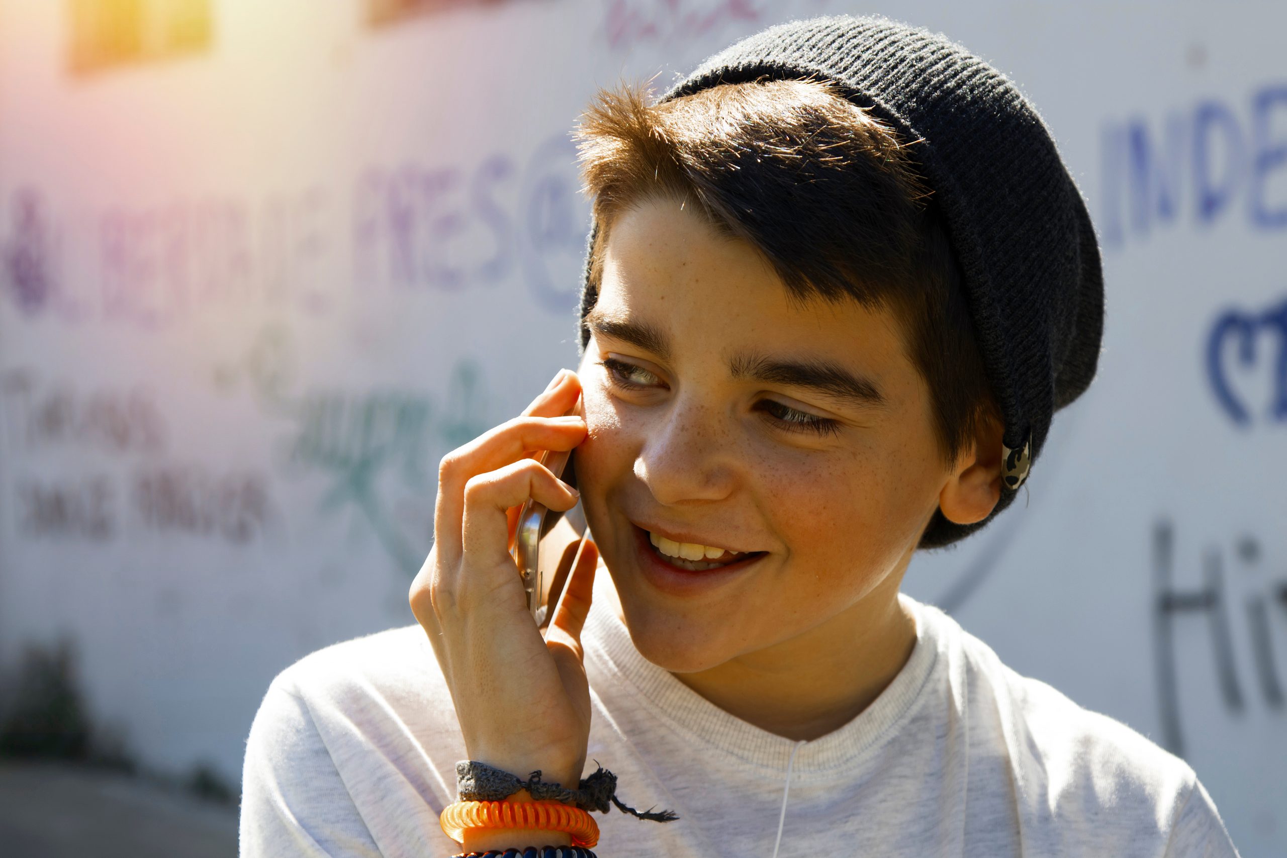 A young person wearing a beanie and a white shirt smiles while discussing one of the "Fifty-Five Creative Uses for Tech" on their smartphone. The background features a wall with colorful graffiti, and sunshine peeks into the scene, adding an artistic backdrop to their conversation.