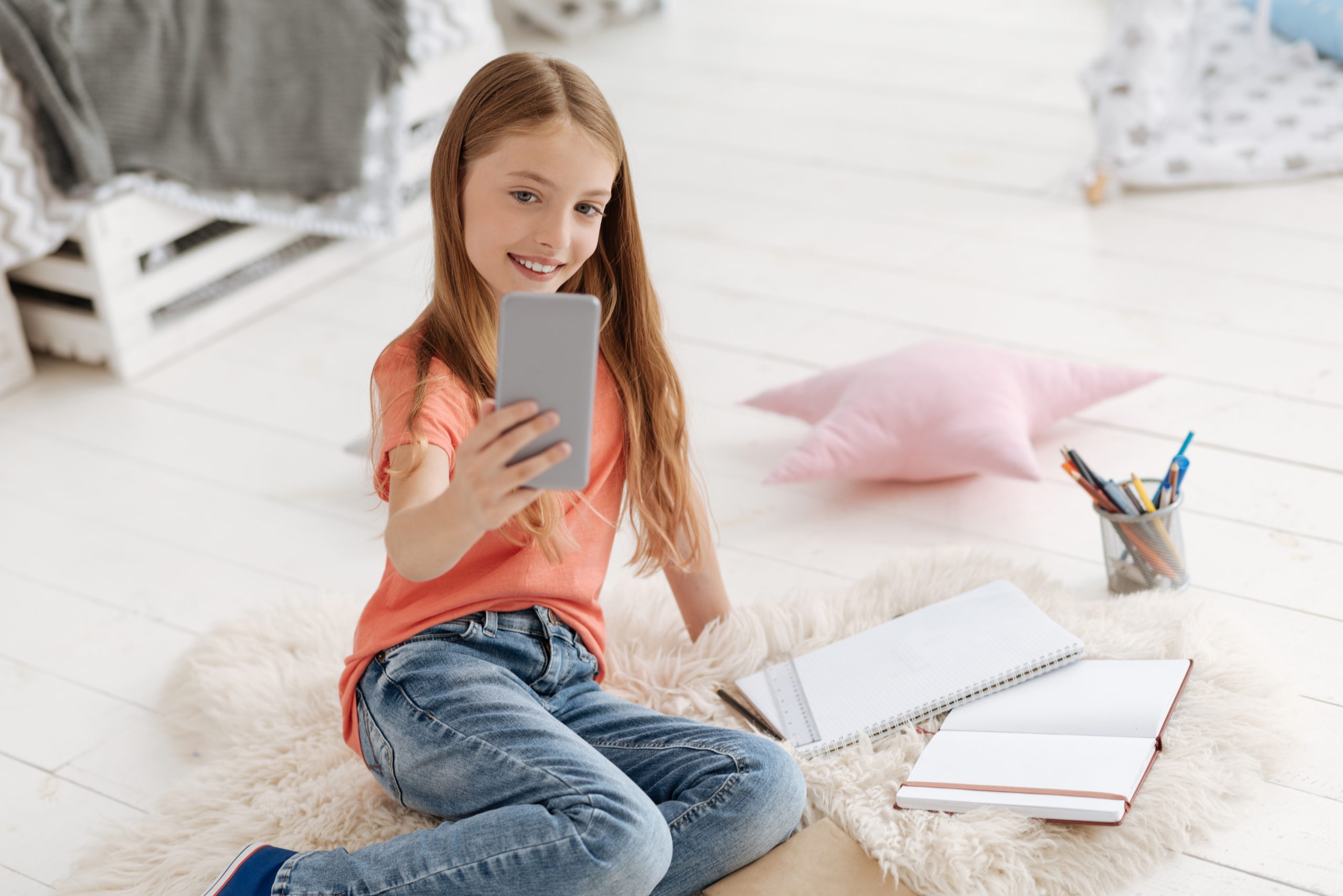 A young girl sits on a fluffy rug, snapping a selfie with her smartphone amidst notebooks, a pencil holder, and a pink pillow. The bright, casually organized setting prompts the question: What if my child gets social media without me knowing?