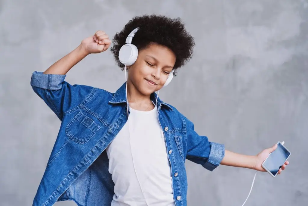 A child in a denim jacket joyfully gets their groove on, dancing with headphones on and holding a smartphone. The scene is set against a light gray wall, perfectly capturing the essence of adding music to your phone.